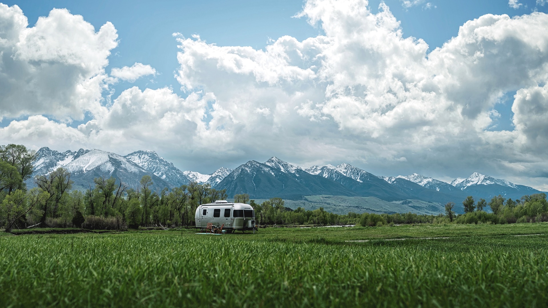 A silver camper is parked on a vibrant green field, with snow-capped mountains and a sky filled with fluffy clouds in the background.