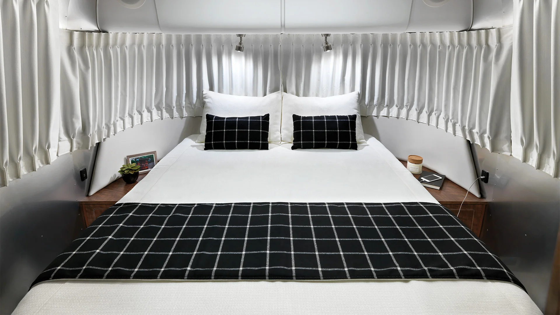 Cozy bed in a vintage trailer with white bedding, black plaid accents, flanked by wooden side tables and white curtains for privacy.