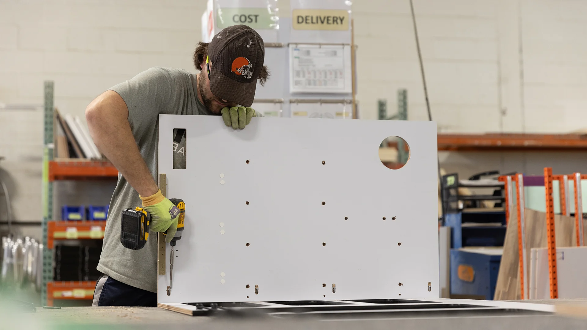 Worker in a workshop uses a drill on a large white panel with holes, wearing a cap, gloves, and safety goggles. Shelves and materials in background.