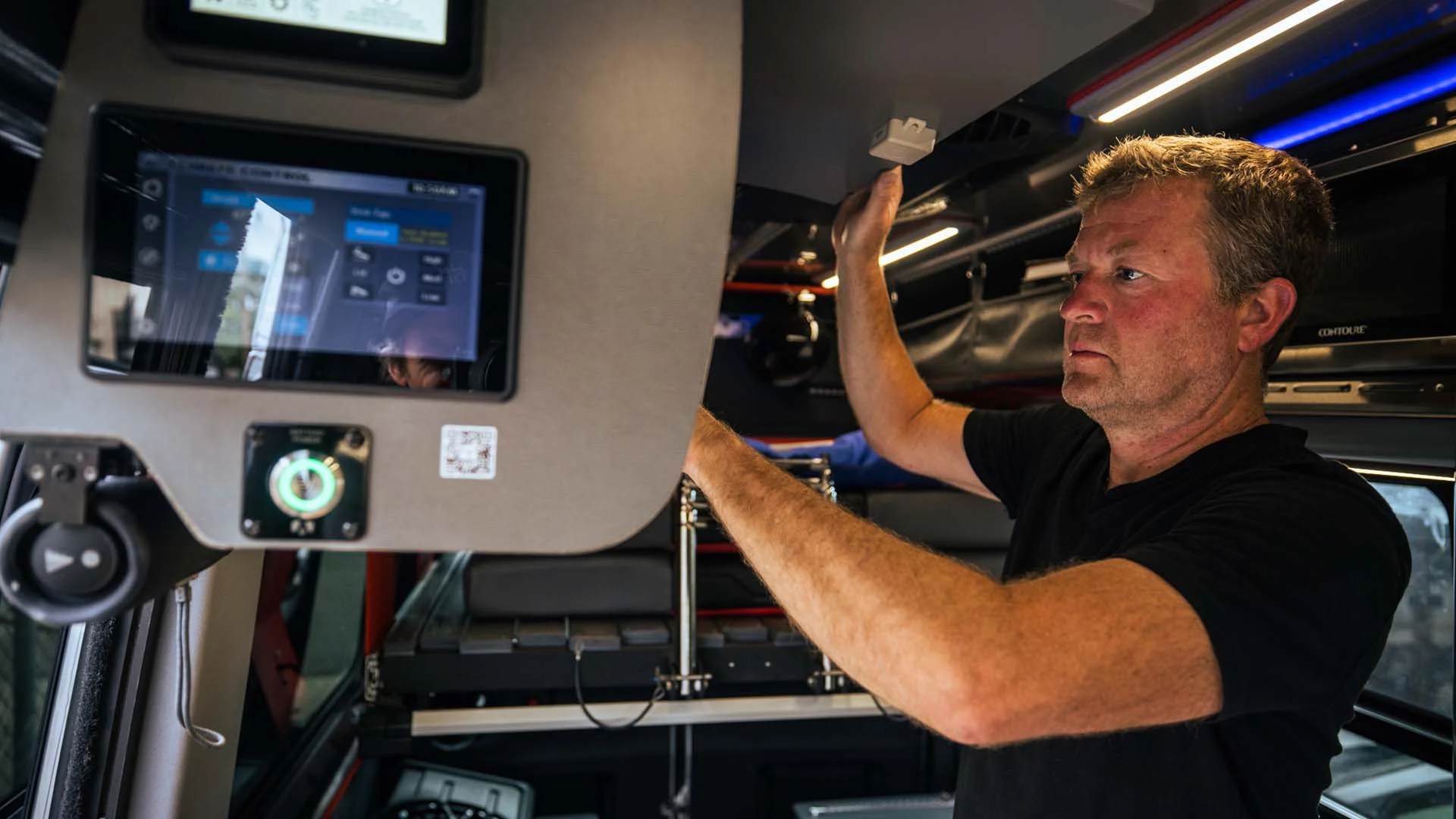 A man adjusts equipment inside a vehicle, with electronic screens and control panels visible around him.