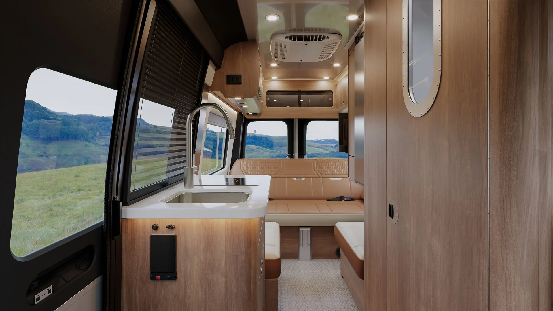 Interior of a modern camper van with wood paneling, a small sink, countertop, seating area, windows showing grassy hills outside, and built-in ceiling lights.