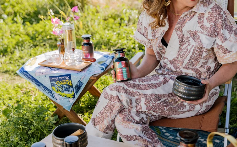 Woman in patterned outfit holds a bottle while sitting outdoors, surrounded by a table with jars, glasses, and flowers on a sunny day.