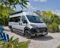 A white camper van parked on a sunny, tropical campsite with palm trees, surrounded by blue chairs and lush greenery.