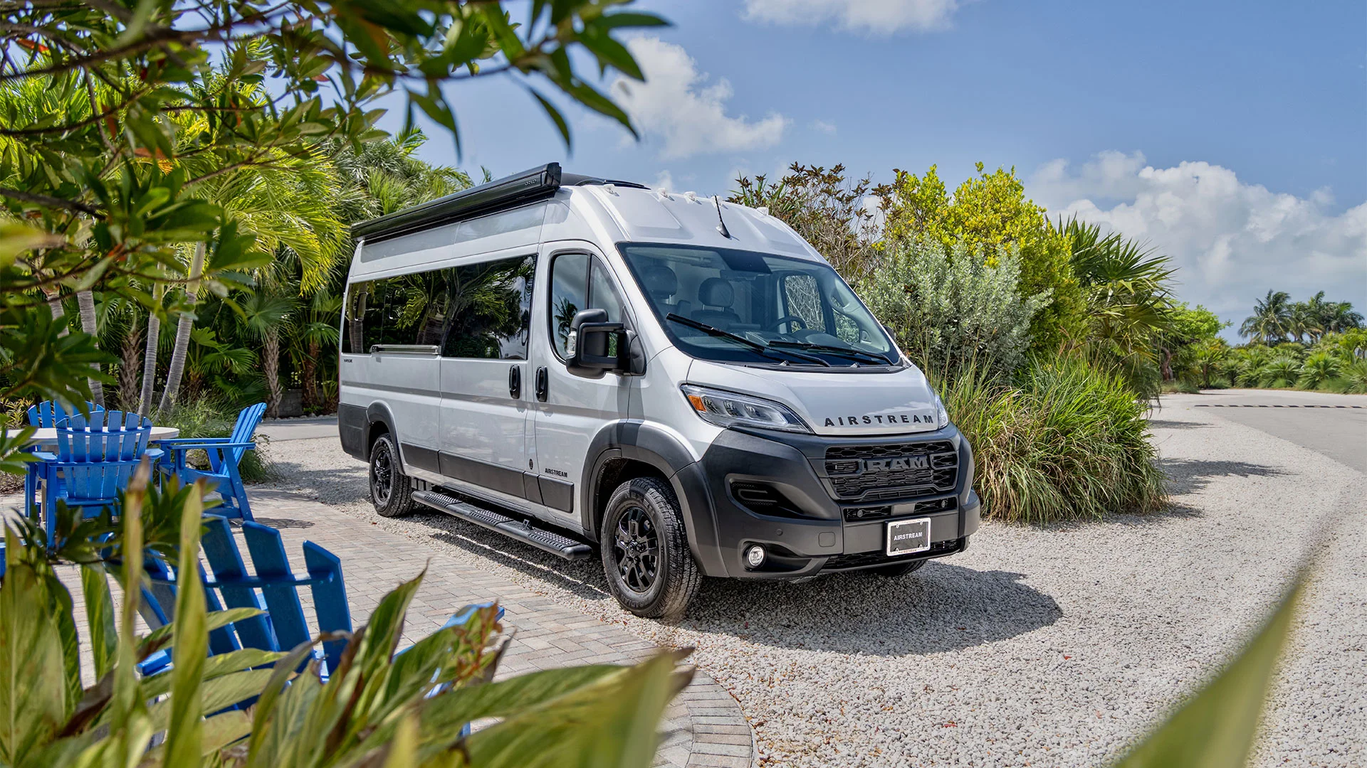 A white camper van parked on a sunny, tropical campsite with palm trees, surrounded by blue chairs and lush greenery.