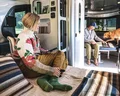 A woman in a floral sweater relaxes with a phone on a bed inside an RV, while a man and a dog are on the seating area.