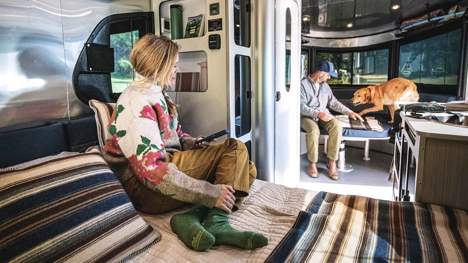 A woman in a floral sweater relaxes with a phone on a bed inside an RV, while a man and a dog are on the seating area.