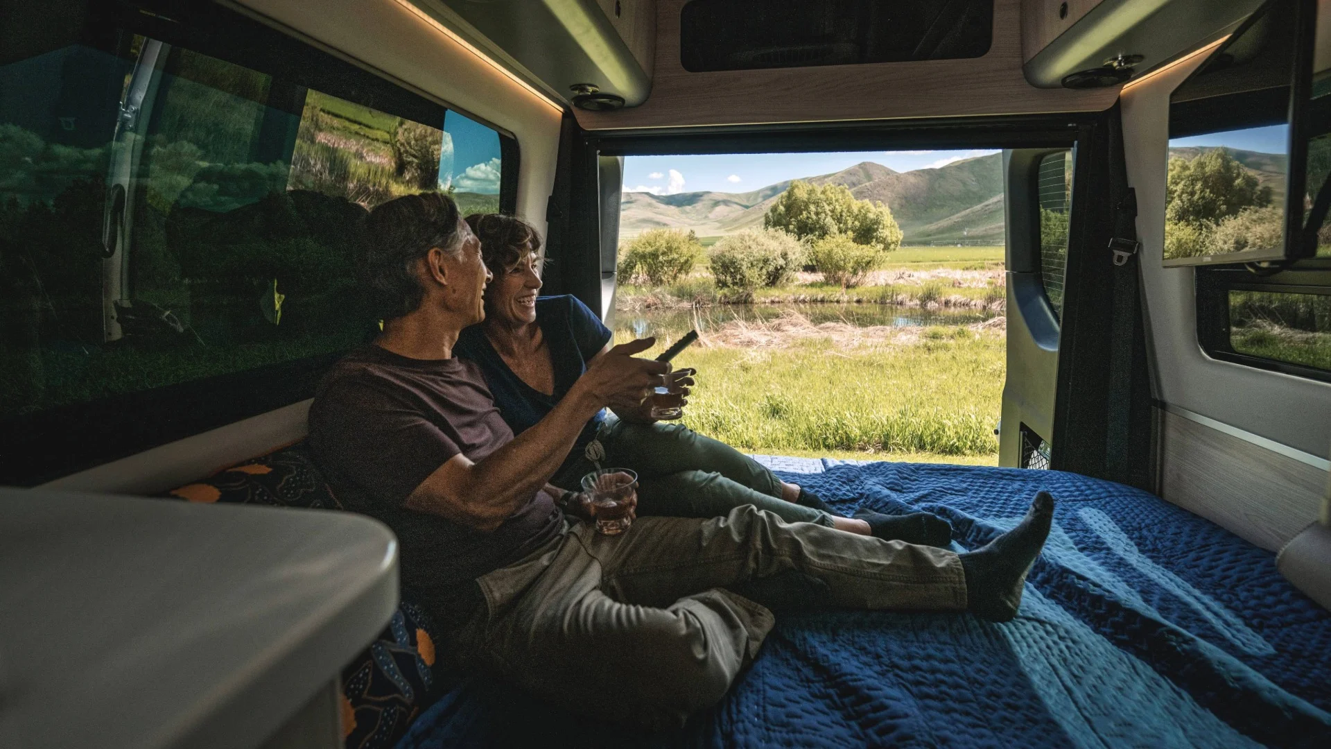 Couple relaxing inside a camper van with open doors, enjoying a scenic view of lush green fields and distant hills.