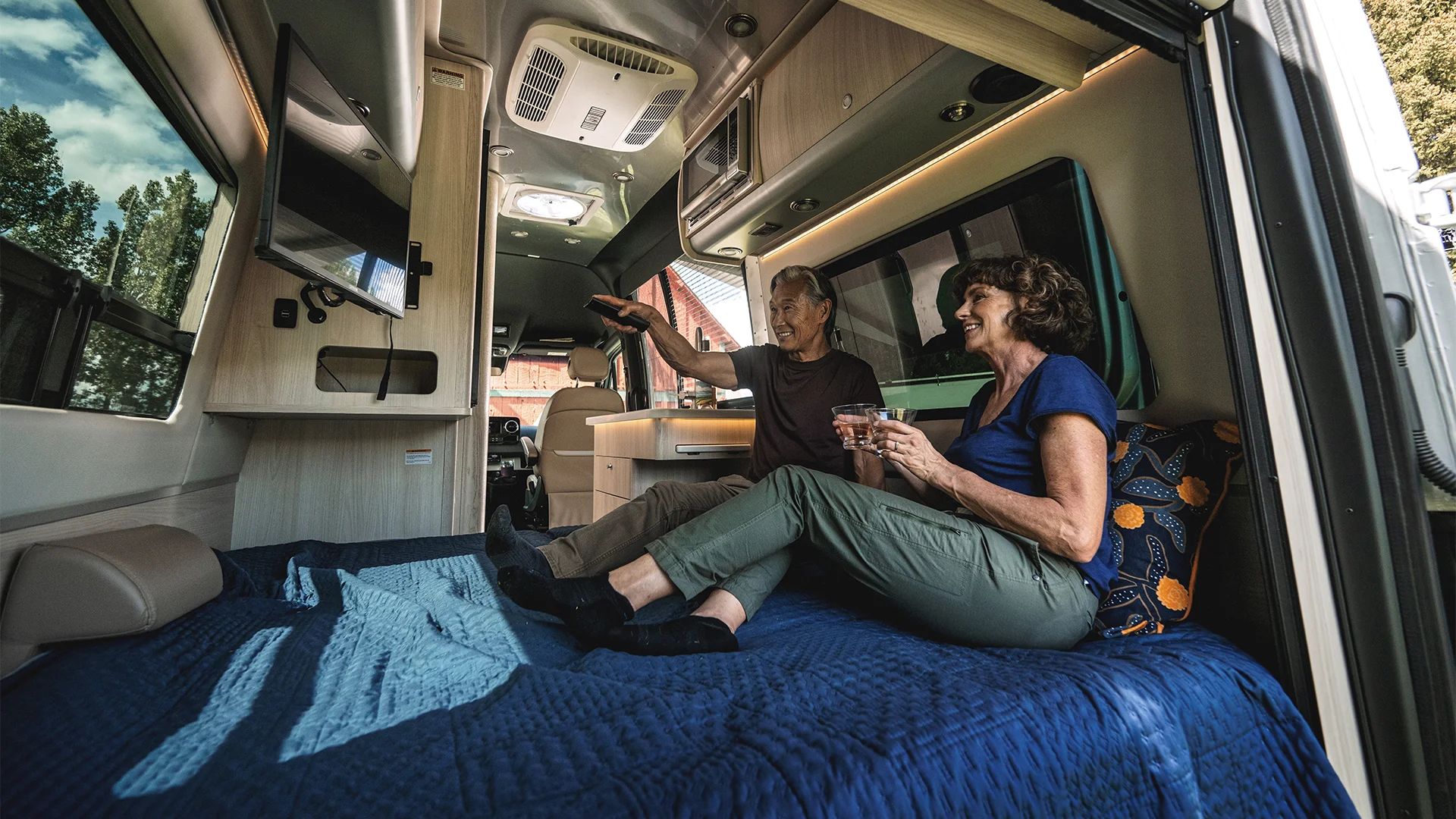 A couple relaxes inside a camper van, sitting on a bed, smiling and holding drinks. They are surrounded by interior amenities and decor.