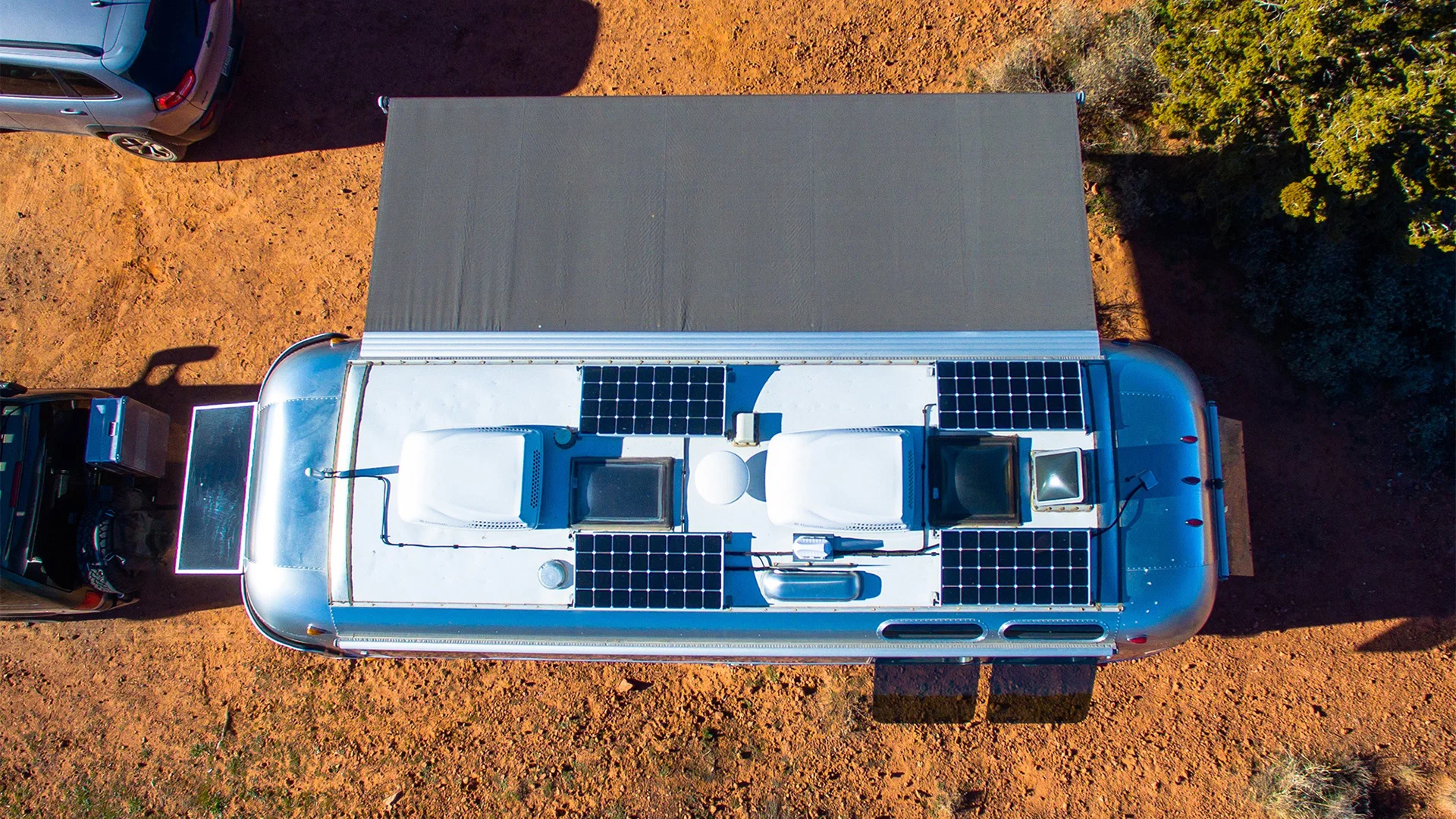 Aerial view of a parked RV with solar panels on the roof, extending an awning on a desert terrain next to a car.