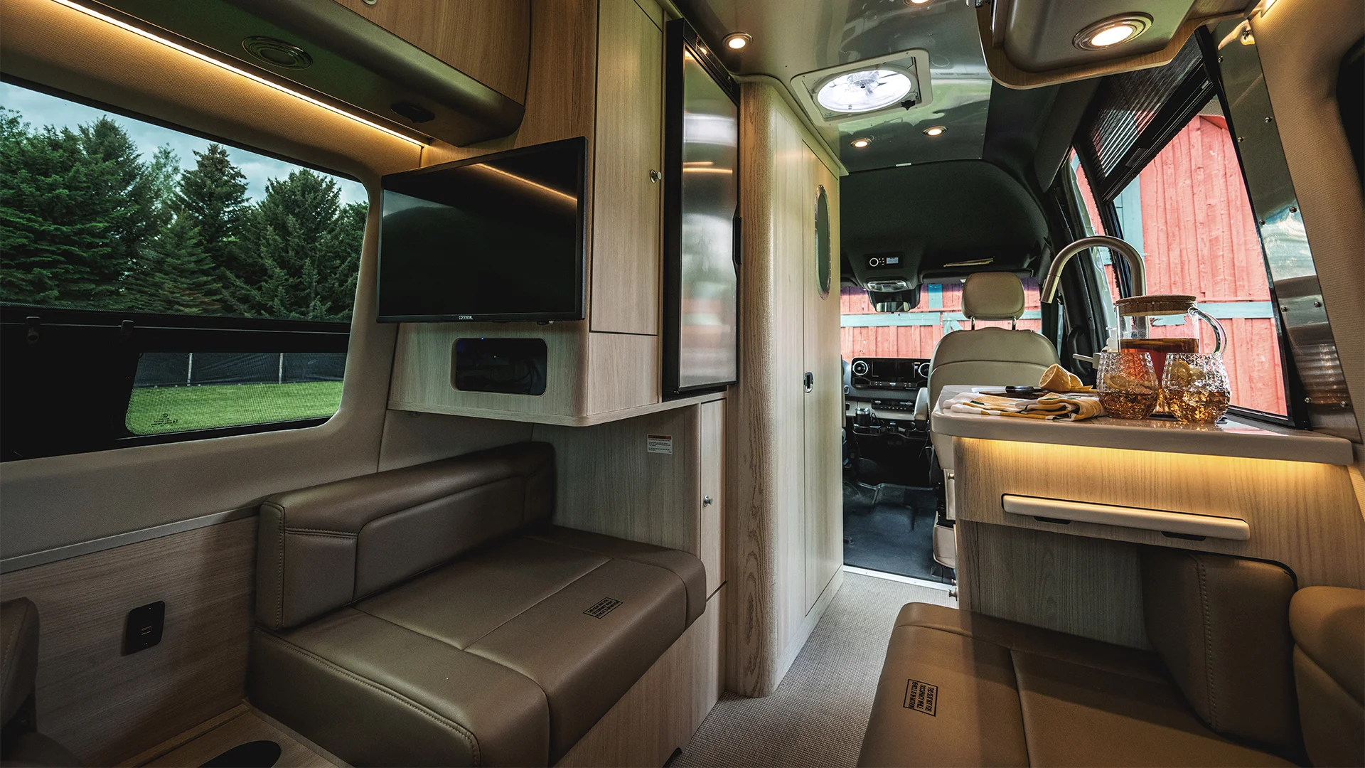 Interior of a luxury RV featuring wood paneling, leather seating, a mounted TV, and a kitchenette with a window view of trees and a red building.