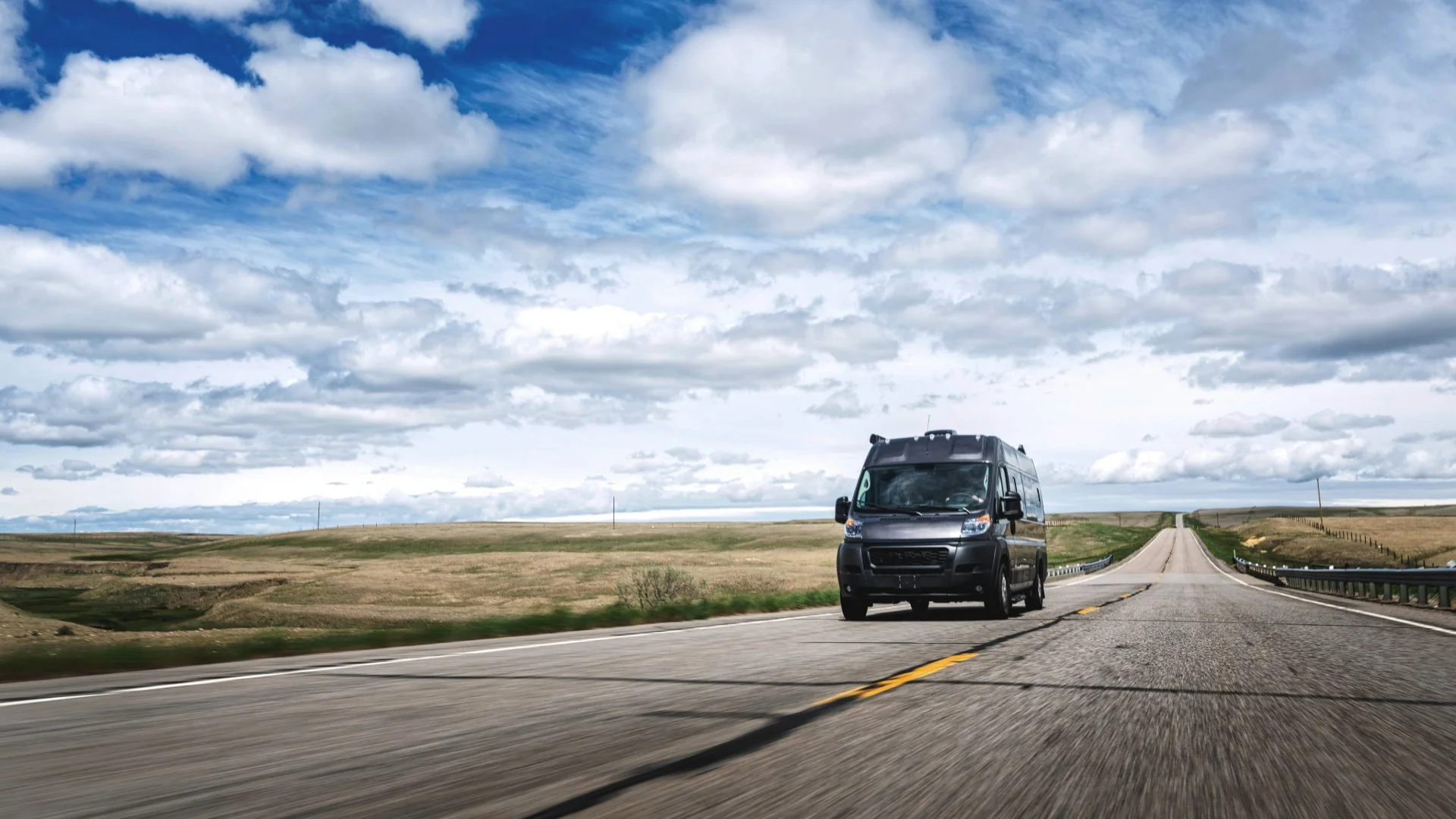 Camper van driving on a long, empty road under a vast, cloudy sky in a rural landscape.