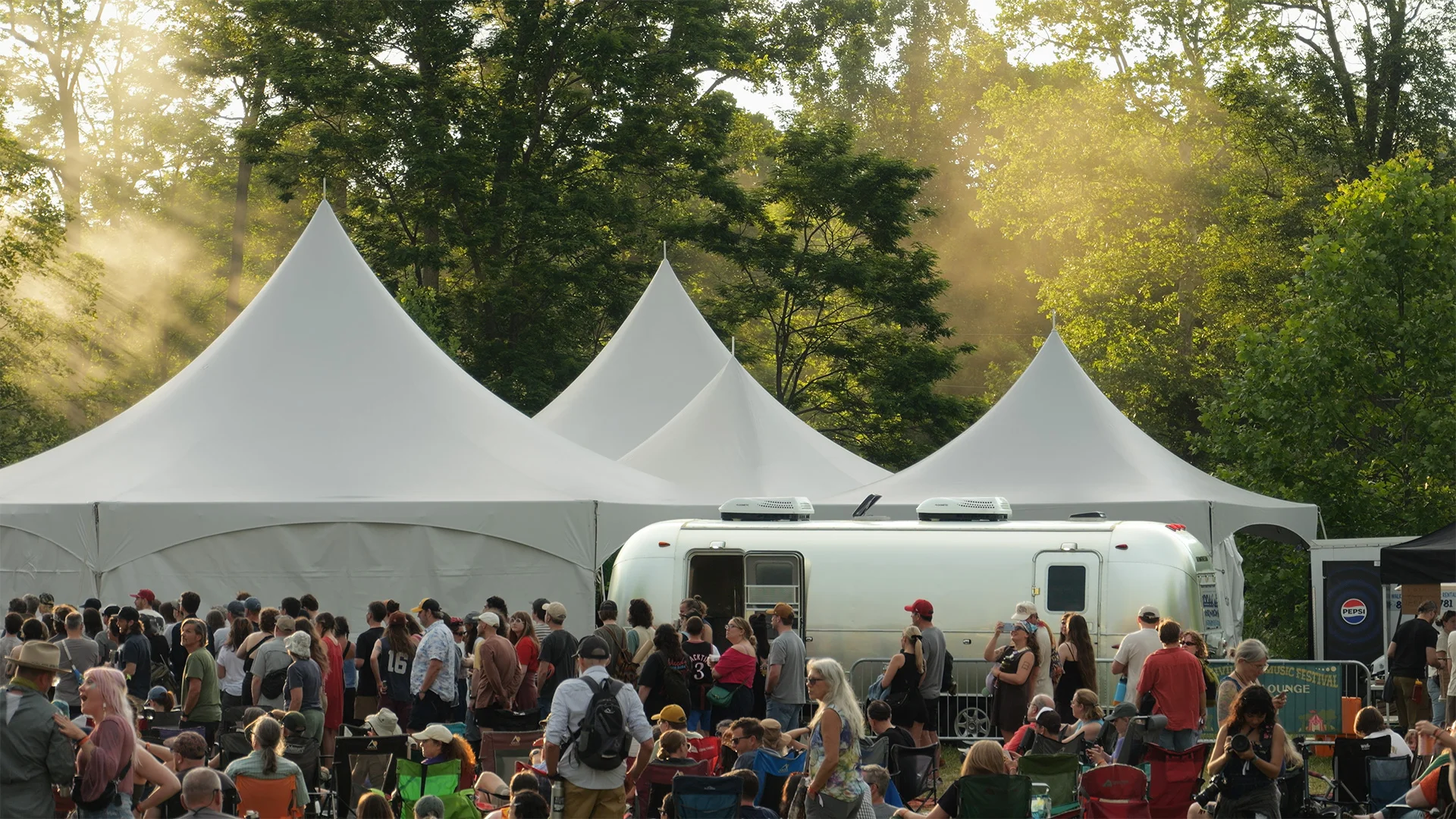 Outdoor festival scene with people gathered around an Airstream bar and beverage trailer in front of large white tents, surrounded by trees and soft sunlight filtering through.