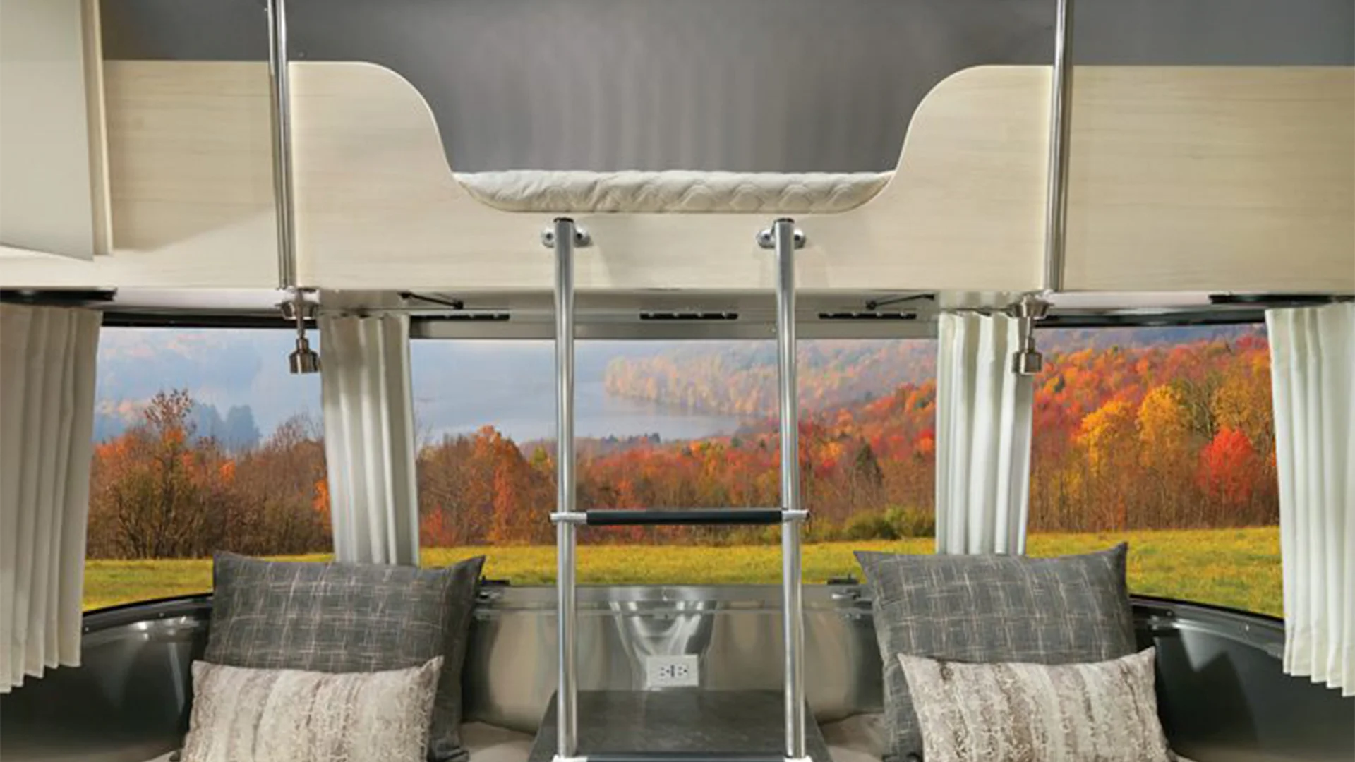 Interior of an RV with a ladder leading to a bunk bed, large windows showcasing vibrant autumn foliage and a serene lake view.