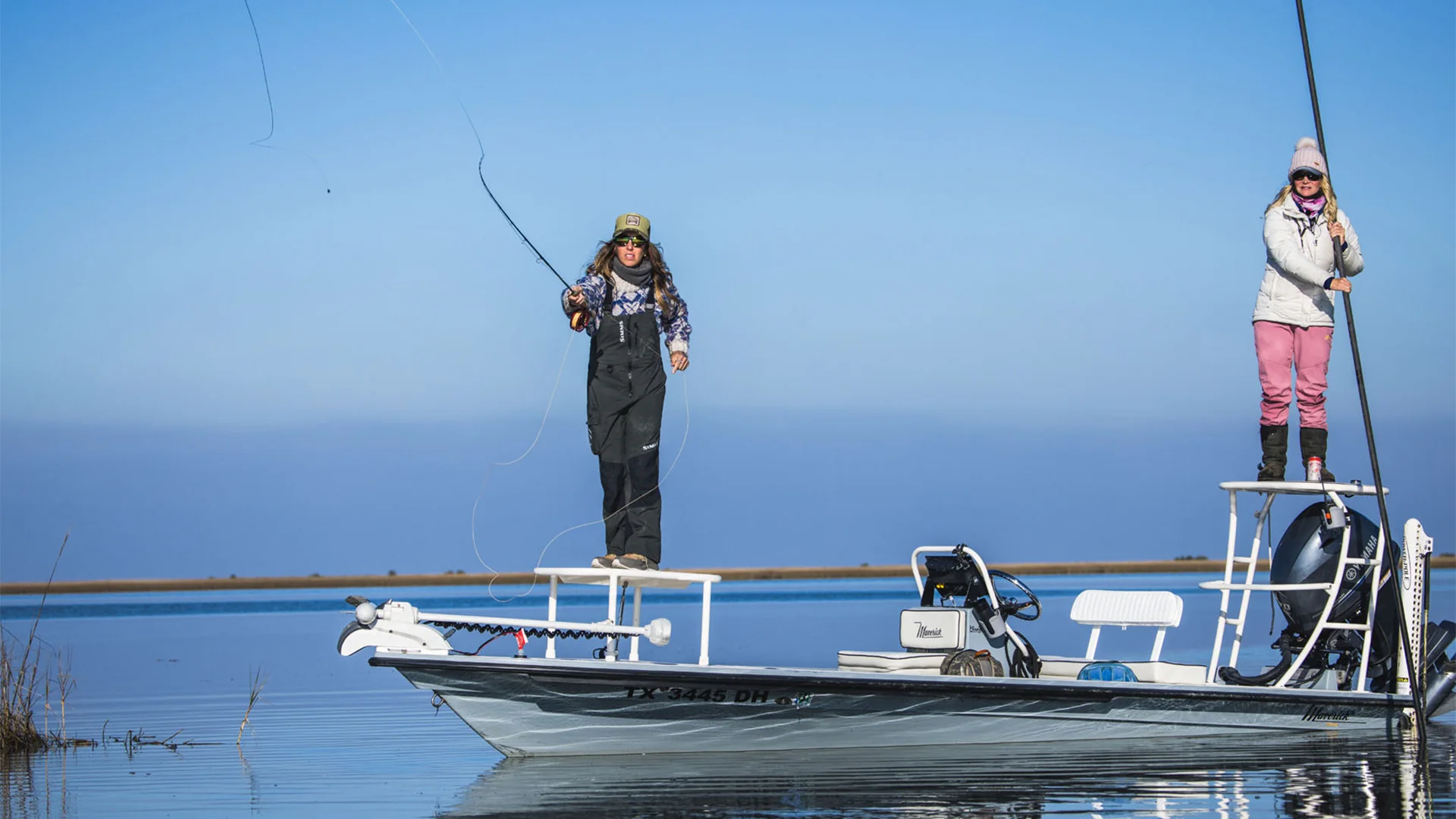 Two people fishing on a small boat in calm water. One person casts a line, while the other stands on a raised platform. Clear sky above.
