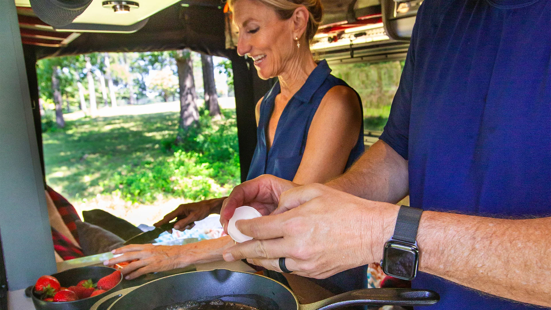 A woman chops vegetables while a man cracks an egg into a skillet in an outdoor kitchen setting.