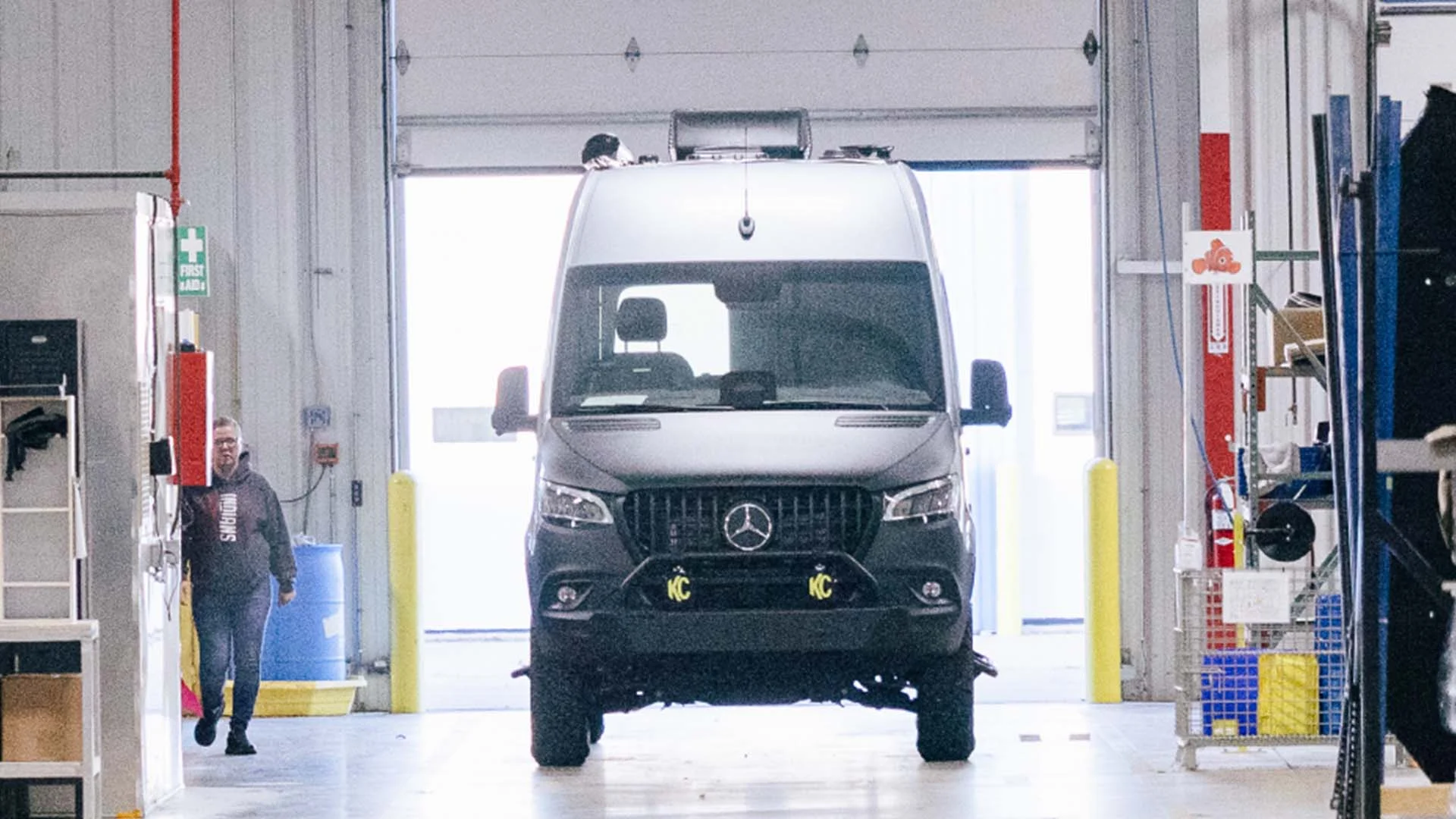 A gray Mercedes van parked inside a spacious garage with industrial equipment and safety signs visible. A person stands nearby.