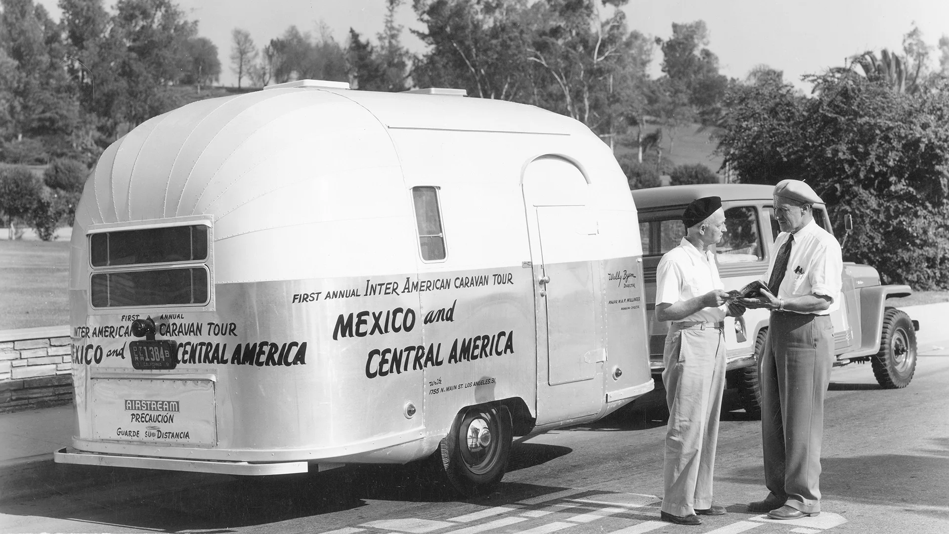 A silver vintage travel trailer labeled Mexico and Central America is hitched to a classic truck. Two men in hats stand beside it, talking and holding papers, with trees in the background.