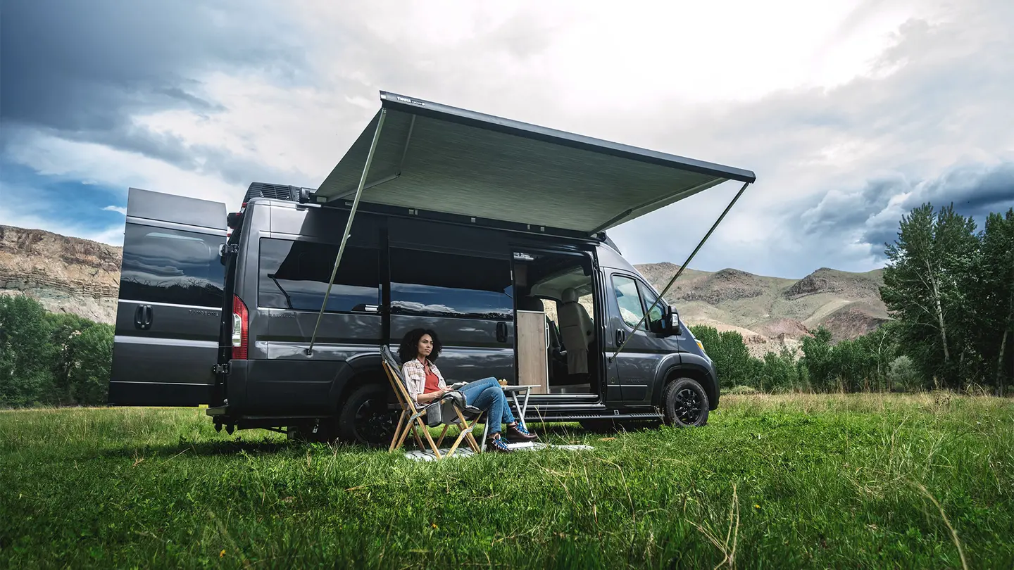 Woman sitting by a camper van with an extended awning, surrounded by mountains and grass, under a cloudy sky.