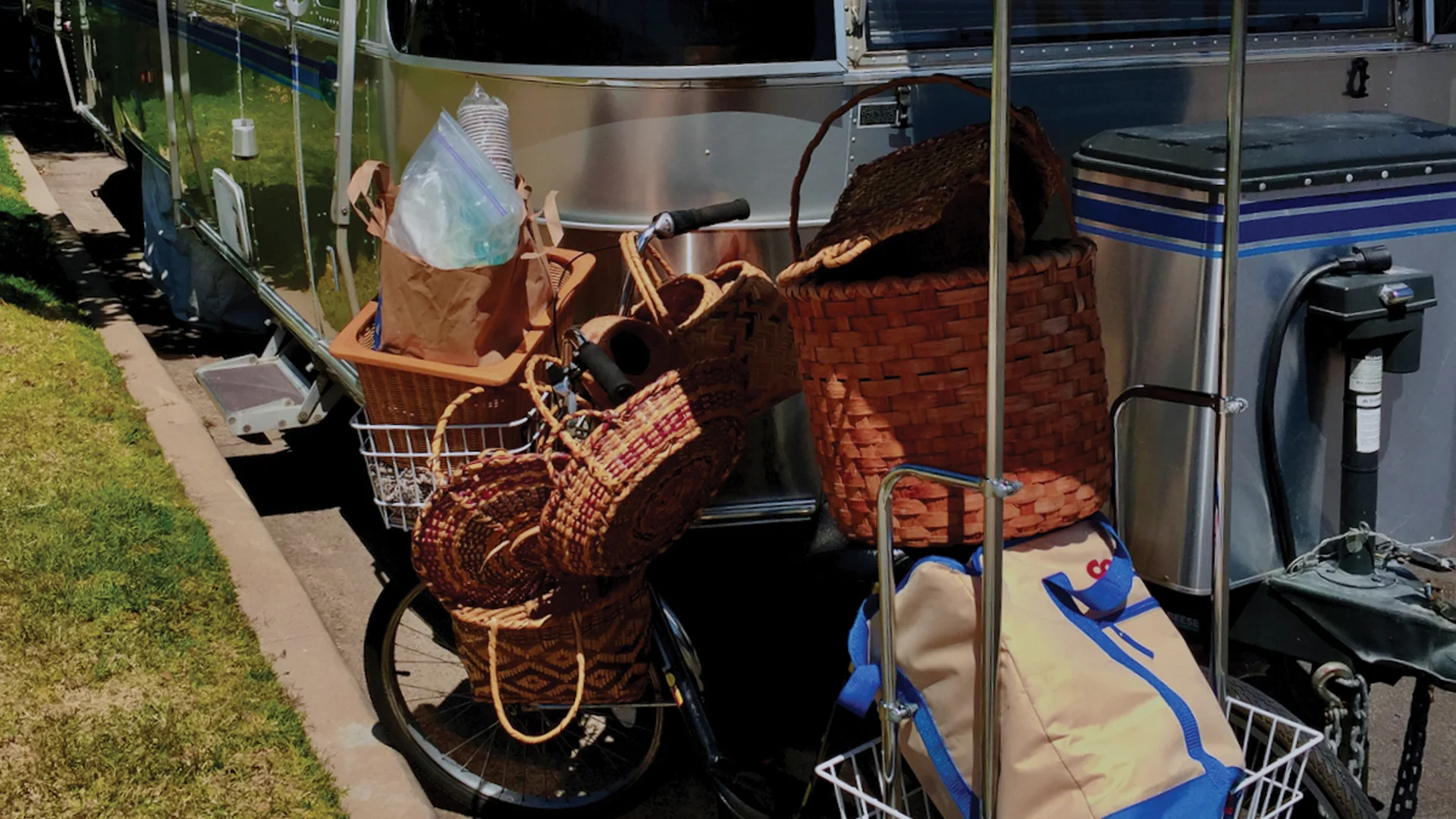 A bicycle loaded with baskets and bags is parked beside a silver camper. The baskets contain various items, and a large woven basket sits on the ground nearby. There is grass and pavement next to the camper.
