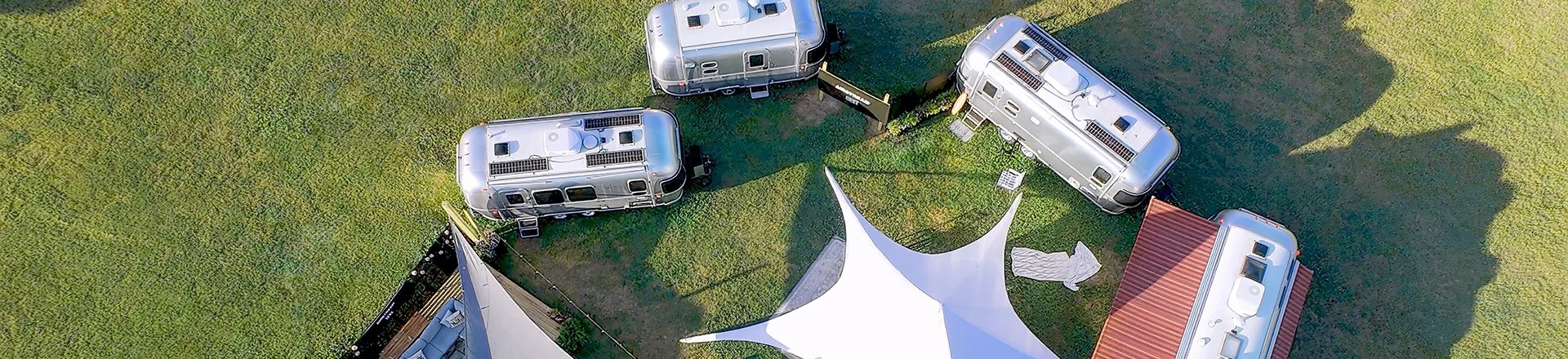 Aerial view of five Airstream trailers arranged in a circular pattern on grass, surrounding a central area with white canopy tents and seating.