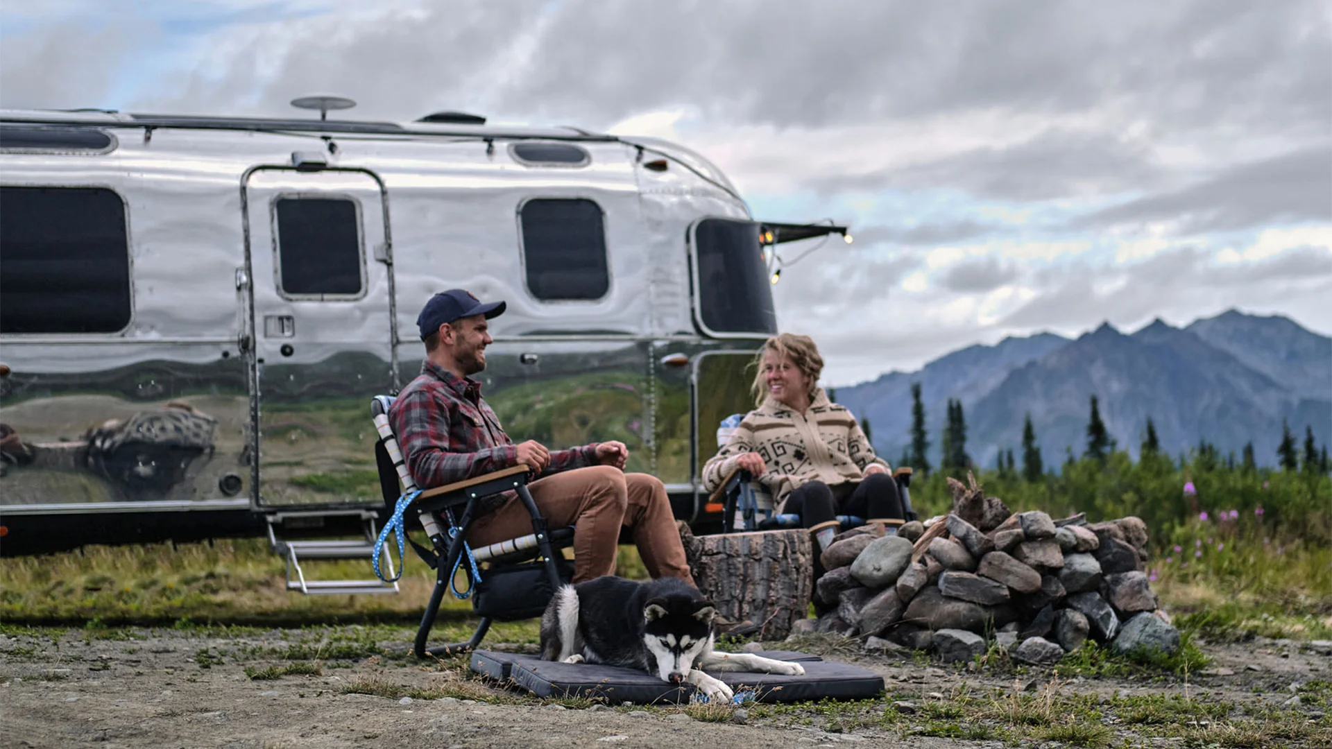 A man and woman sit by a campfire next to an Airstream trailer with a dog resting nearby, set against a mountain backdrop.