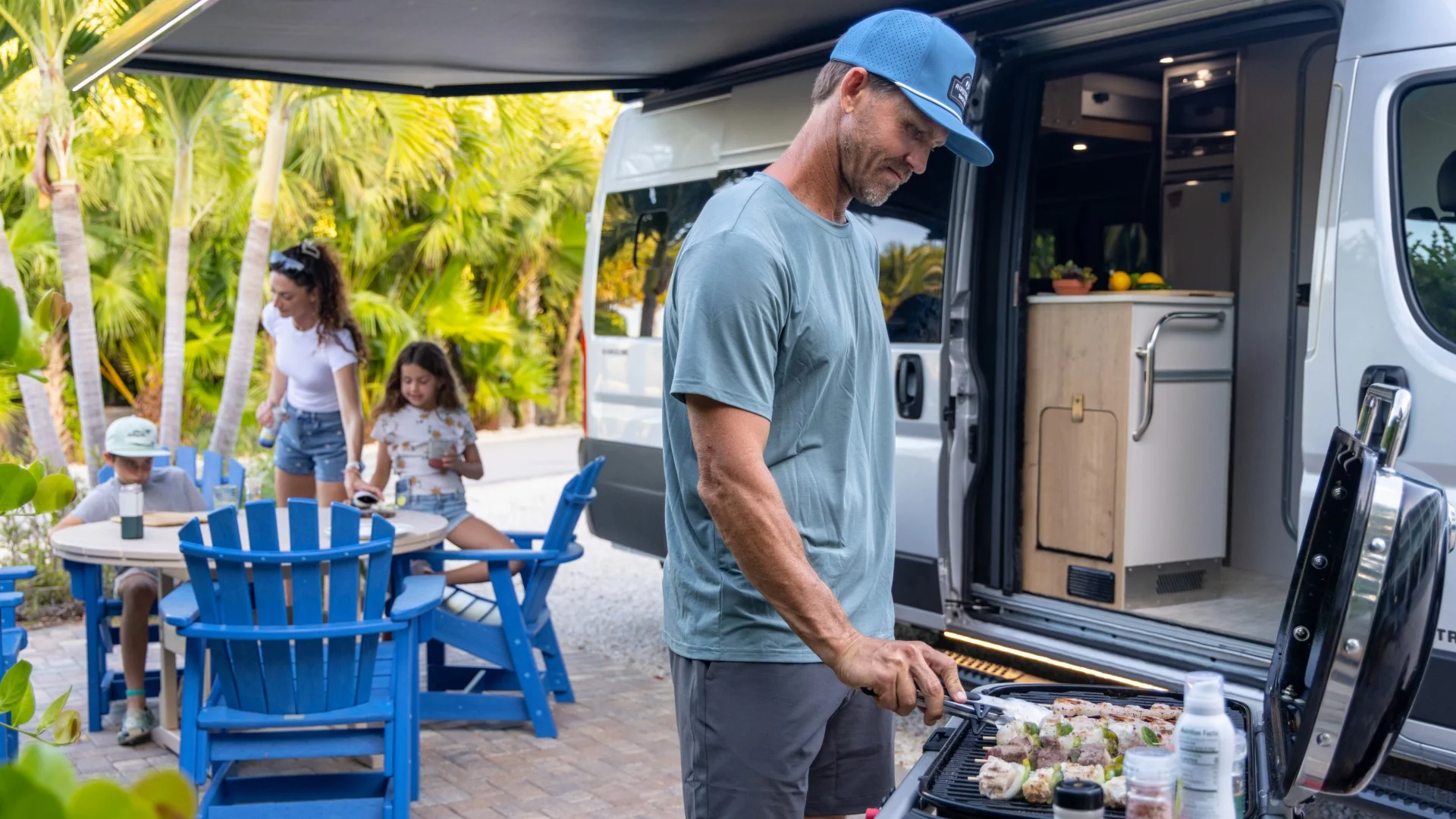 Man grilling kebabs outside a camper van, with a woman and two children at a blue table, surrounded by palm trees.