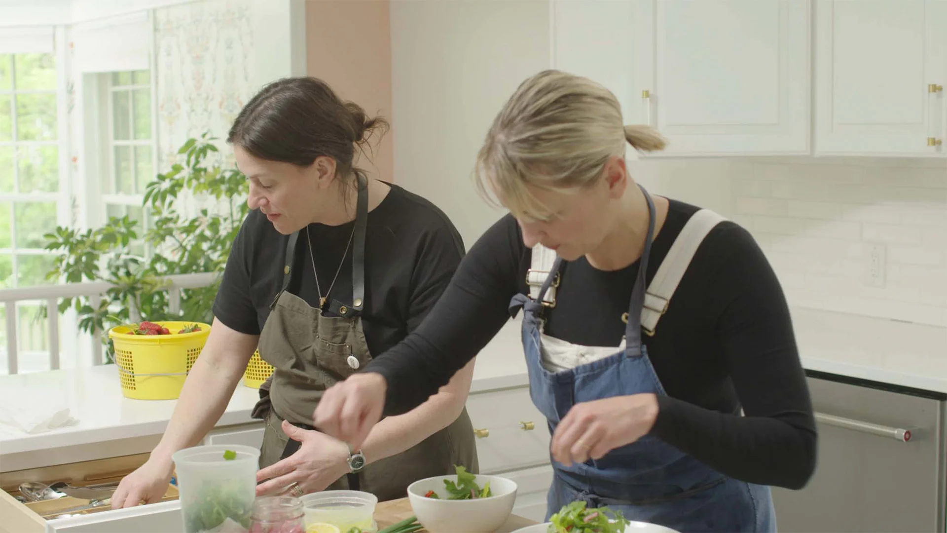 Two women in aprons prepare food in a bright kitchen, focused on chopping and arranging ingredients in bowls.