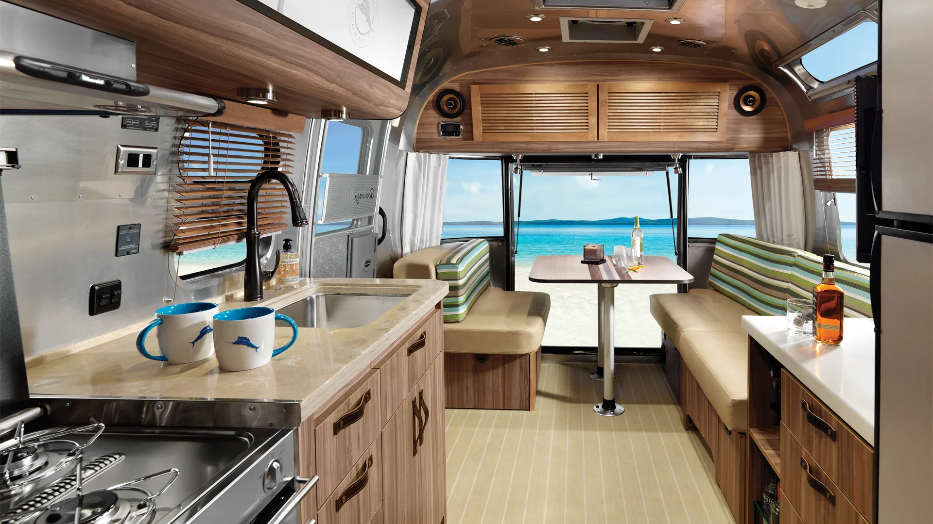 Interior of a modern camper van with wooden cabinets, a compact kitchen, two mugs on the counter, and a dining area with striped seats, looking out to a beach and blue ocean through large rear windows.