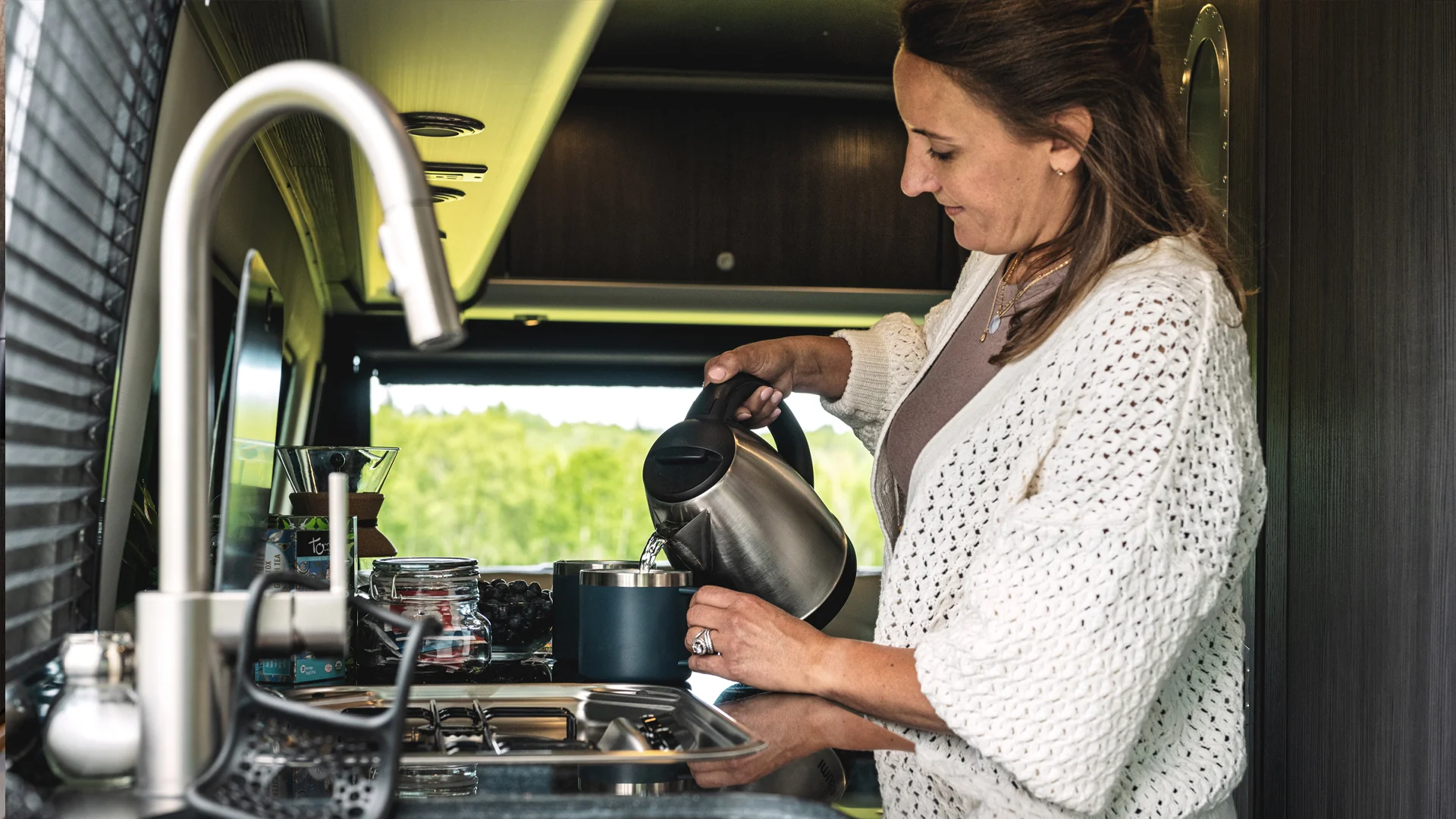 Woman pouring water from a kettle into a pot in a modern kitchen with a sink and various kitchen items around her.
