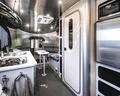 Interior of a modern camper van with a small kitchen, stove, sink, refrigerator, and a seating area with a table. The design features sleek metallic finishes and compact, efficient use of space.