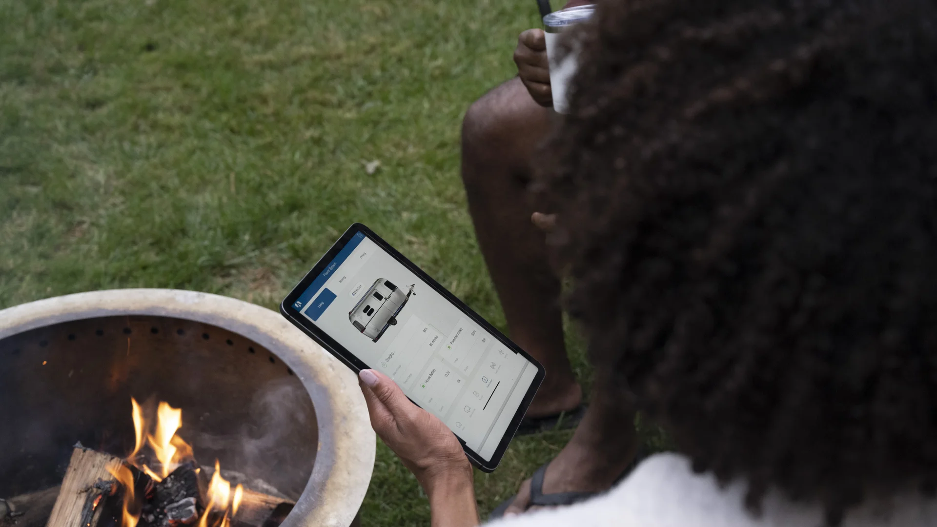 Person holding a tablet near a small outdoor fire pit on grass, viewing an image of a camper van on the screen.