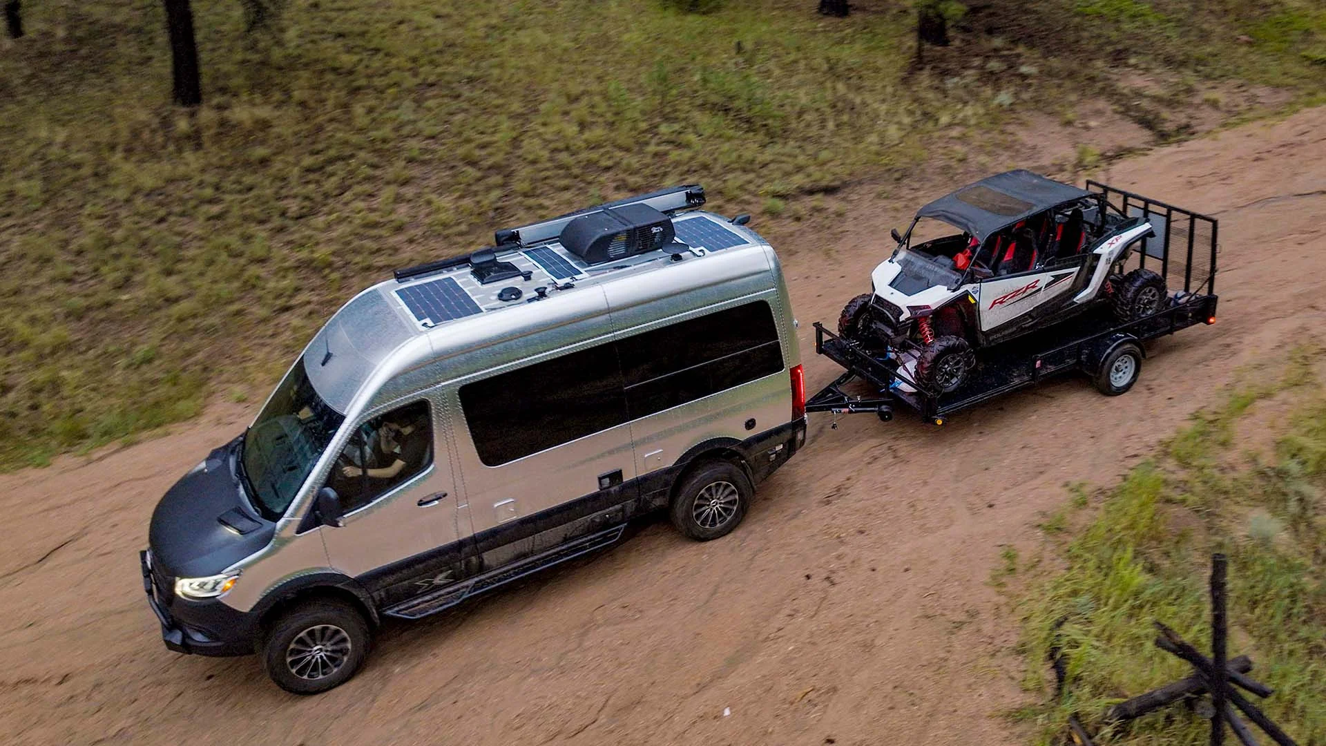 A camper van towing an off-road vehicle on a trailer along a sandy path, surrounded by grass and trees.