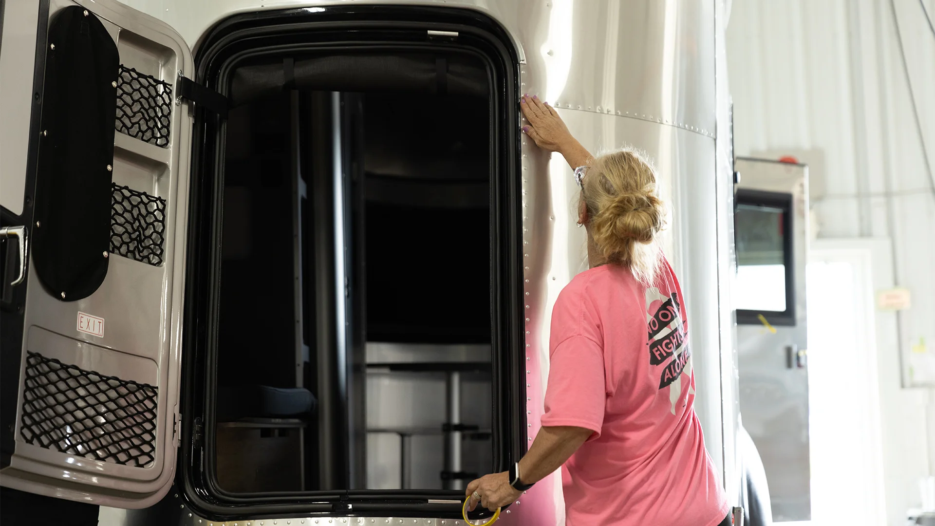 Person in a pink shirt cleans the exterior of an open trailer inside a garage, with light filtering through a window in the background.