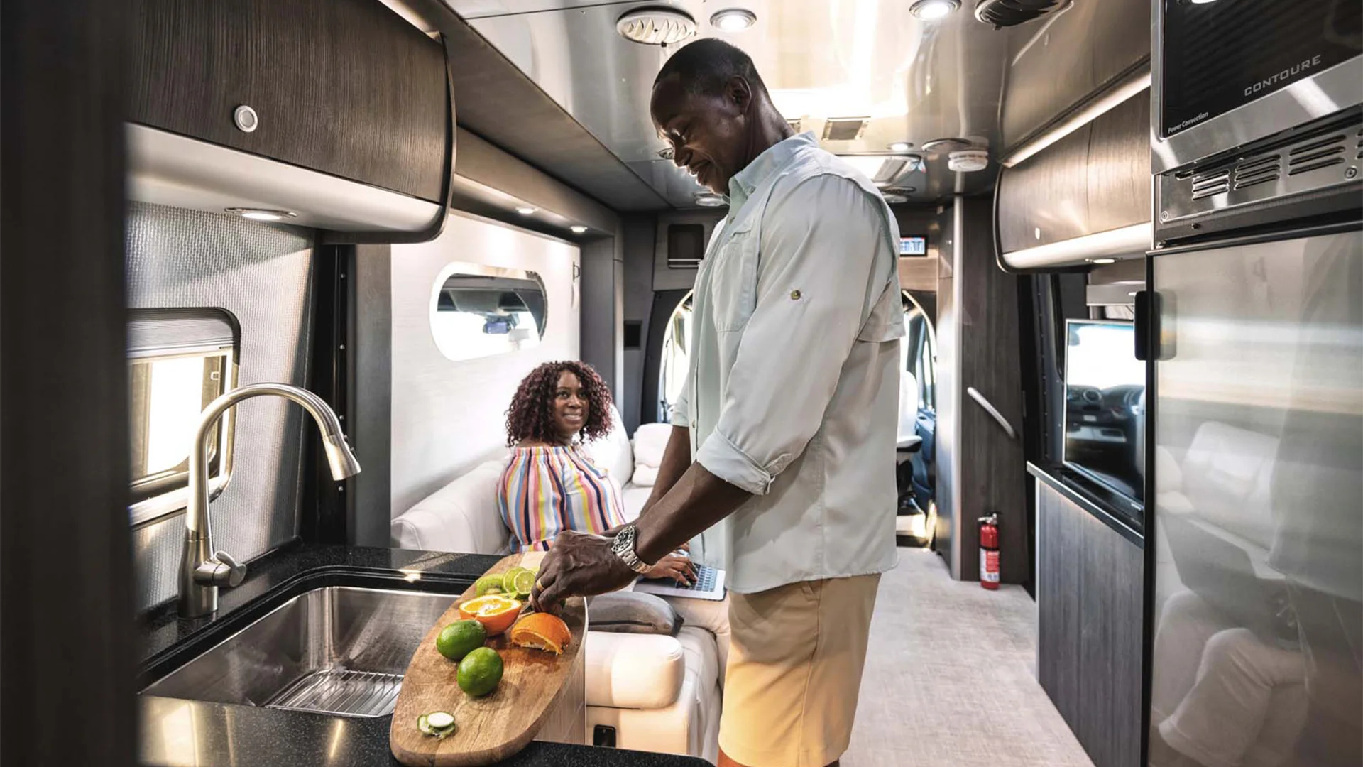 Man slicing citrus fruits on a cutting board in a modern RV kitchen, with a woman sitting in the background, looking at a laptop.