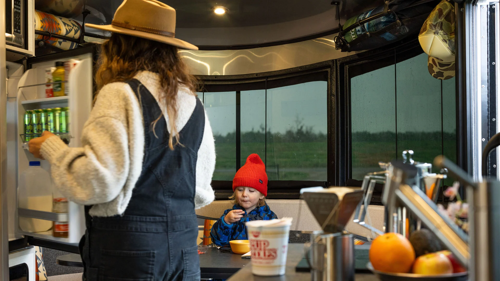 A woman in a hat opens a fridge inside a camper while a child in a red beanie eats at the table. The camper’s windows show a rainy landscape outside. Fruit and a cup of noodles sit on the kitchen counter.