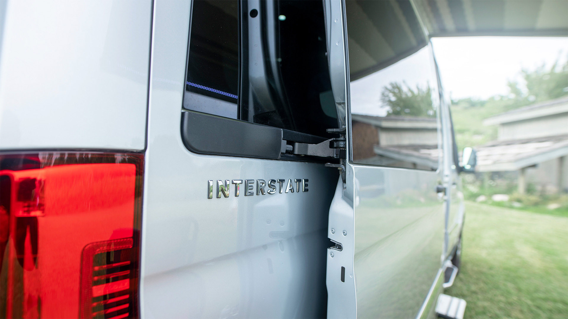 Close-up of a silver van with "INTERSTATE" logo, rear door open, and red taillight visible. Background shows greenery and buildings.