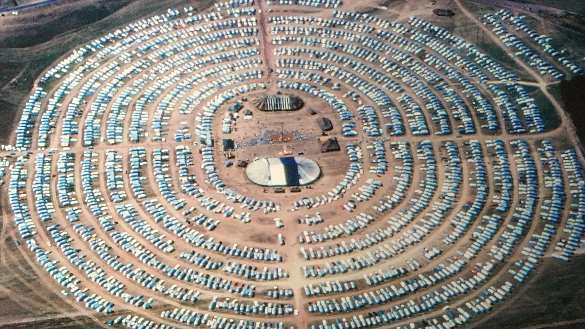 Aerial view of a large circular campsite with hundreds of tents arranged in concentric rings around a central open area on dry, brown ground.