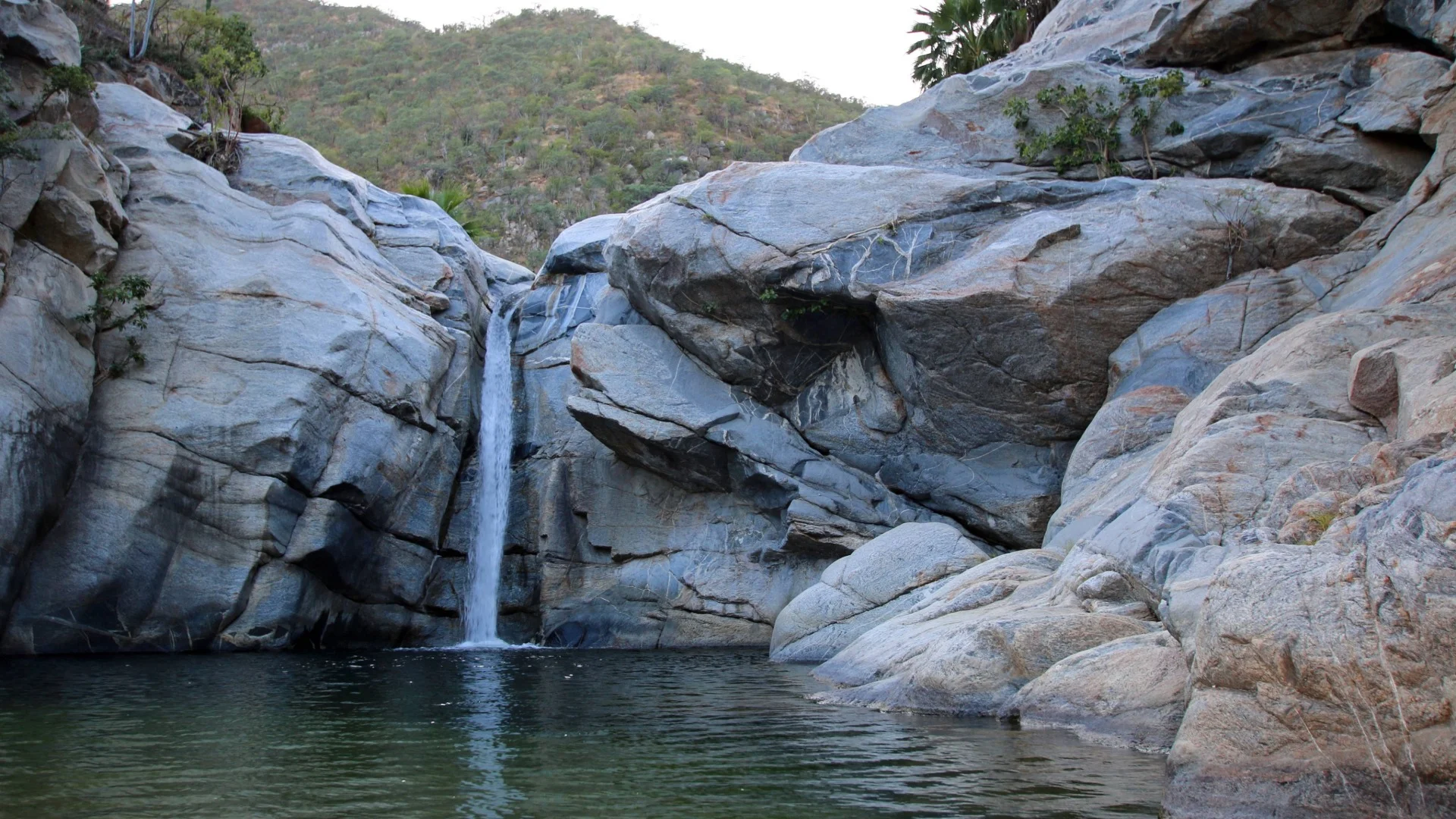 Small waterfall cascading over gray rocks into a calm pool, surrounded by rugged terrain with sparse vegetation.