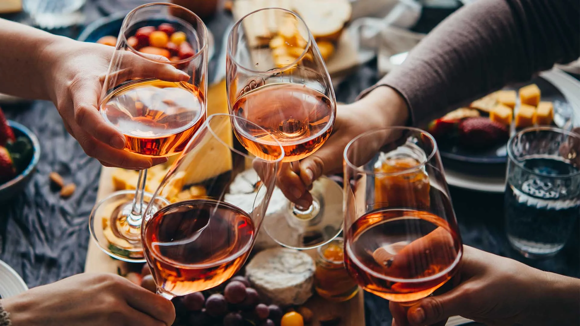 Four people clink glasses of rosé wine over a table with cheese, grapes, and snacks, creating a celebratory and convivial atmosphere.