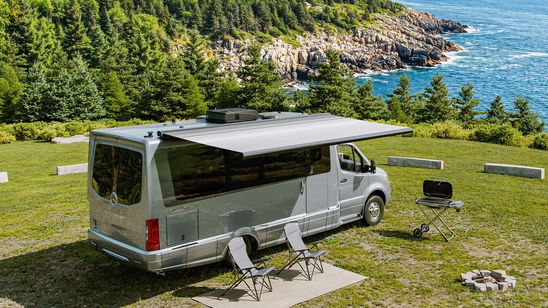 A camper van with an extended awning is parked near a coastal landscape, with two chairs and a grill set up on a grassy area.