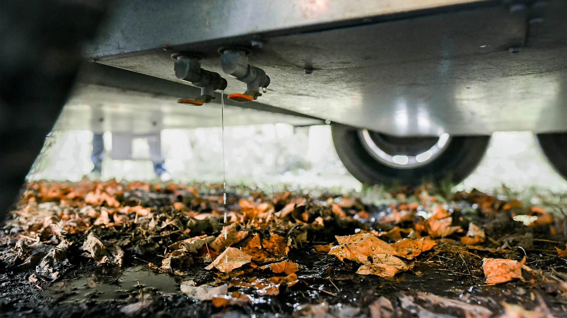 Water drips from a valve under a vehicle or trailer, onto wet ground covered with fallen autumn leaves, with one wheel visible in the background.