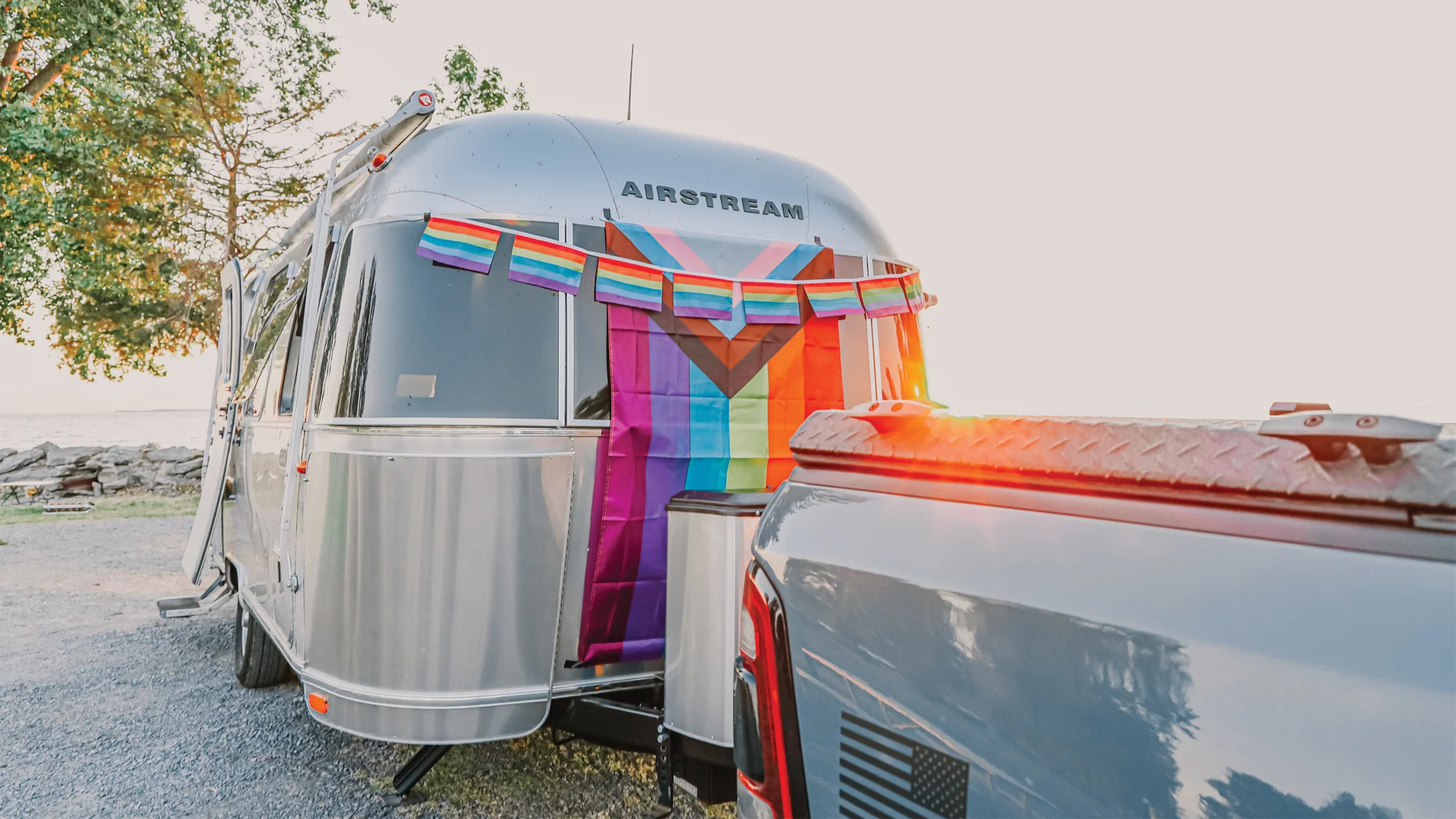 A silver Airstream trailer decorated with rainbow Pride and Progress Pride flags is parked near a body of water, with a pickup truck hitched in front and sunlight shining from the right.