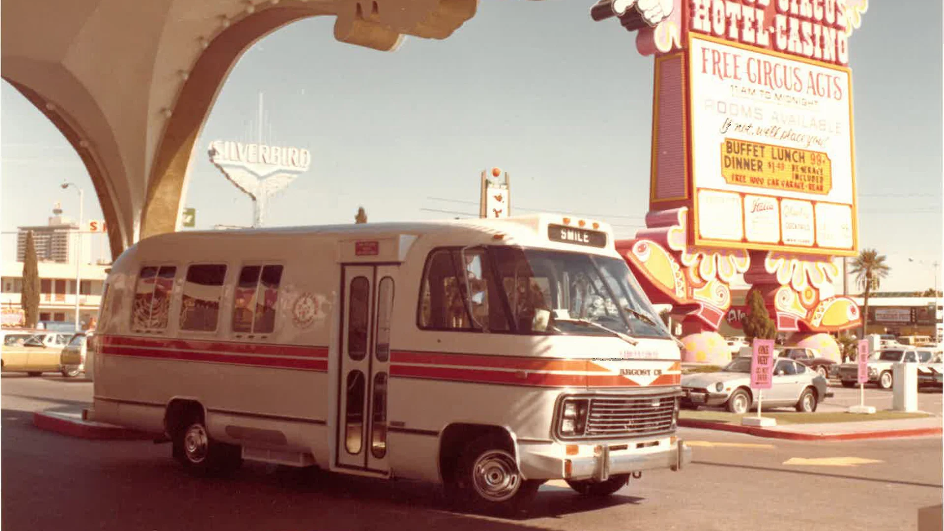 A vintage RV with red stripes parked under a large arch in front of the Circus Circus Hotel and Casino in Las Vegas, with 1970s-era cars and colorful signs in the background.