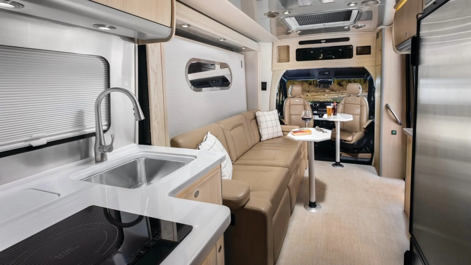Modern RV interior with a kitchenette, beige sofa, small dining table, and swivel chairs. Cozy and functional space for travel.