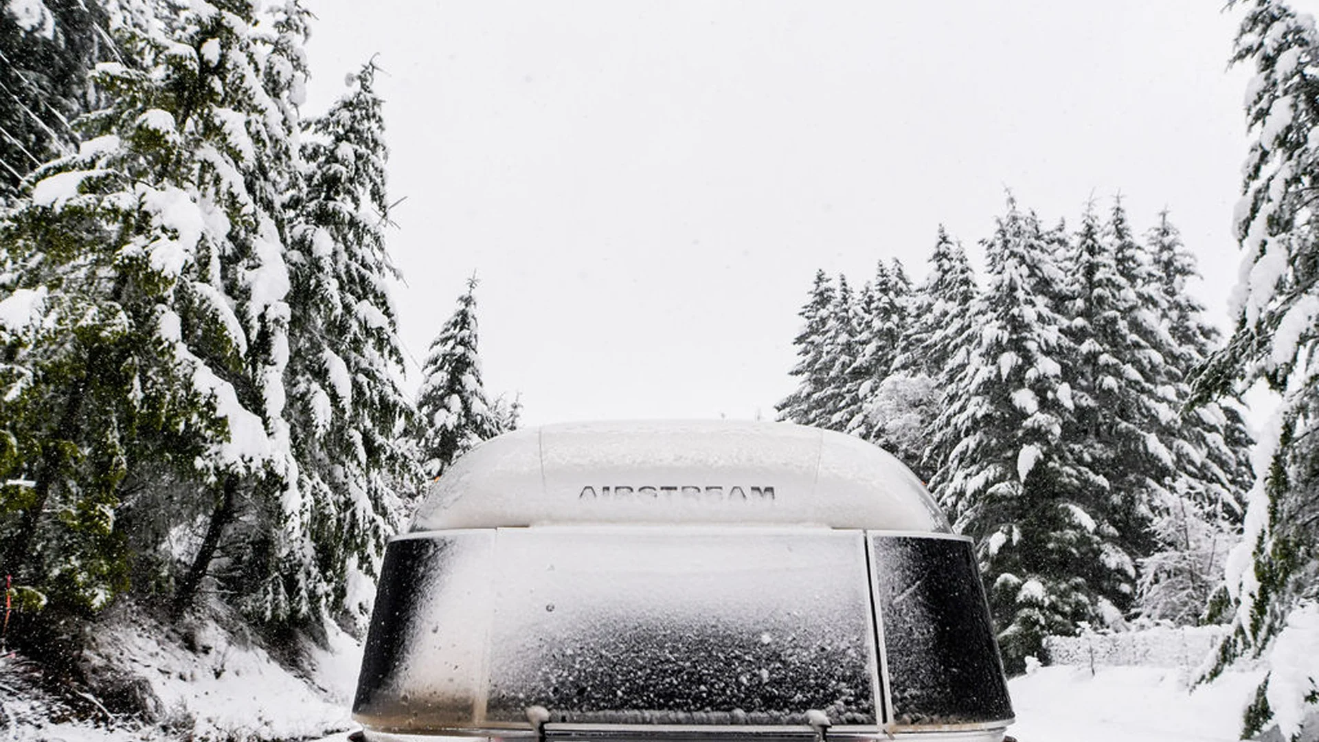 A silver Airstream trailer is parked on a snowy road, surrounded by snow-covered evergreen trees under a gray winter sky.