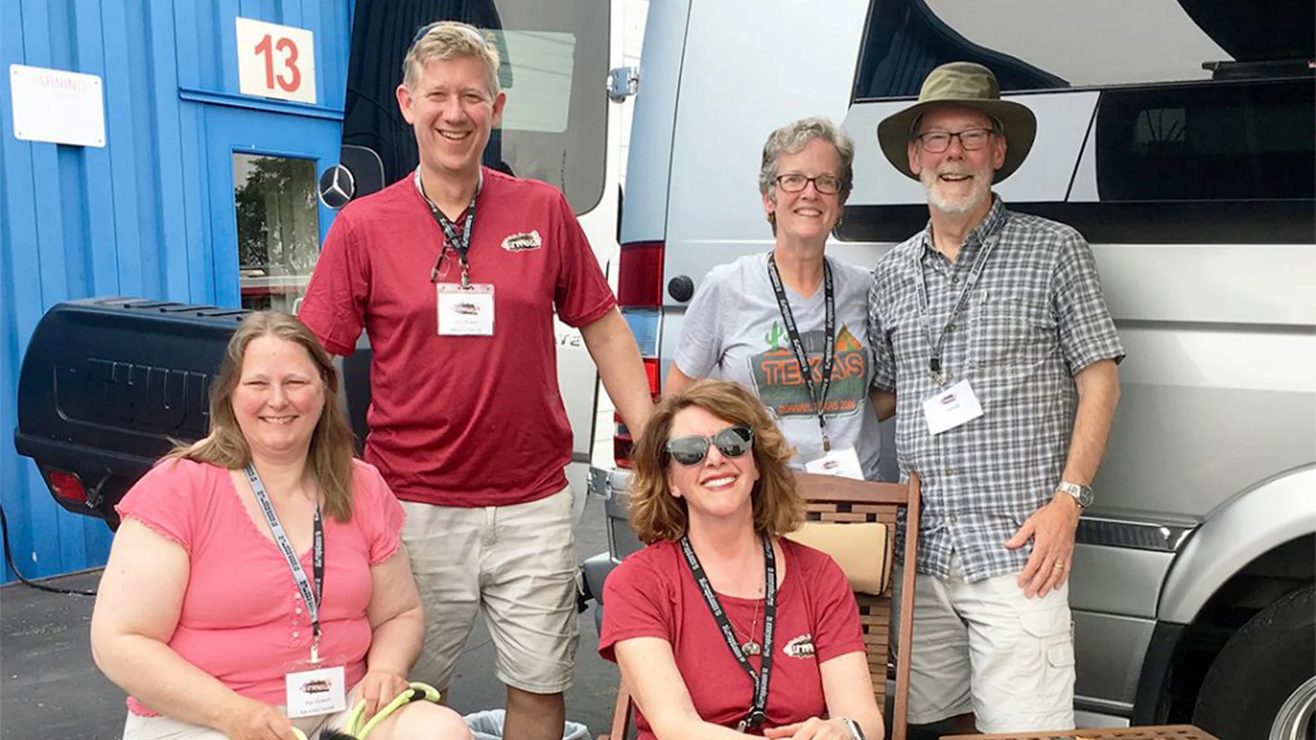 Five people smiling outdoors, in front of a van, wearing name badges. Two are seated, three are standing. Blue building in the background.