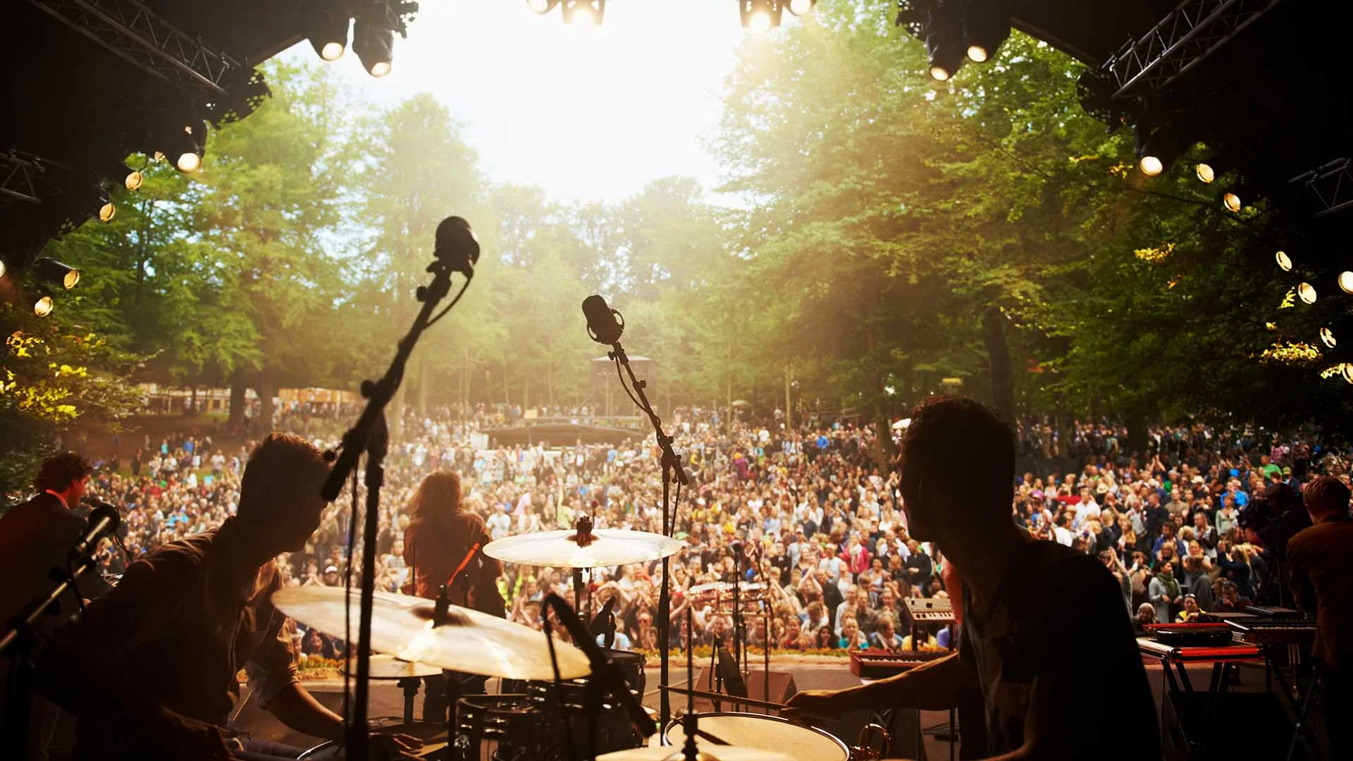 Musicians perform on an outdoor stage, seen from behind the drum set, as a large crowd gathers in front of them among green trees in daylight. Stage lights shine above, illuminating the lively scene.