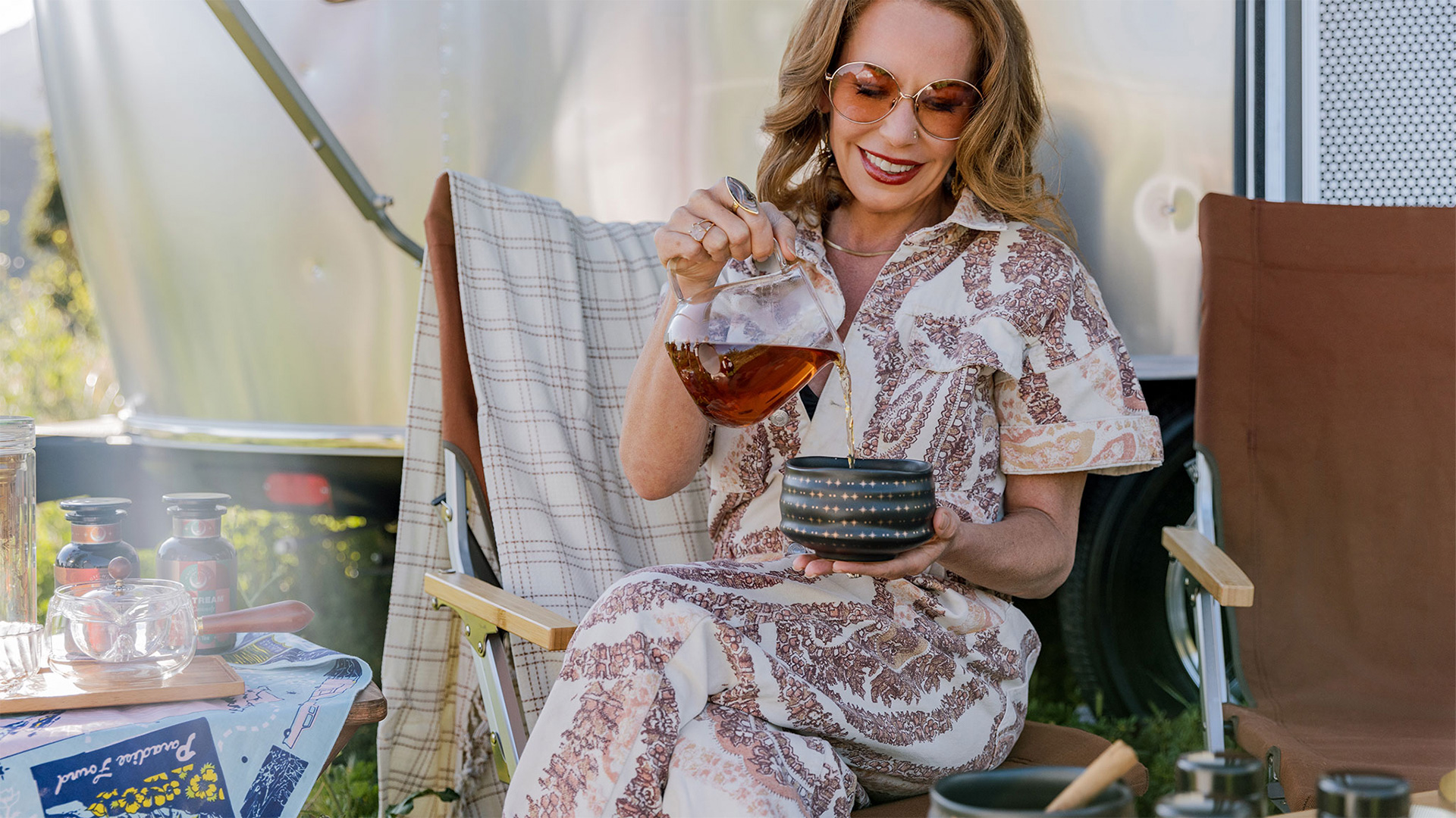 Woman in patterned outfit pours tea into a cup while seated outdoors near a trailer, with a picnic setup around her.