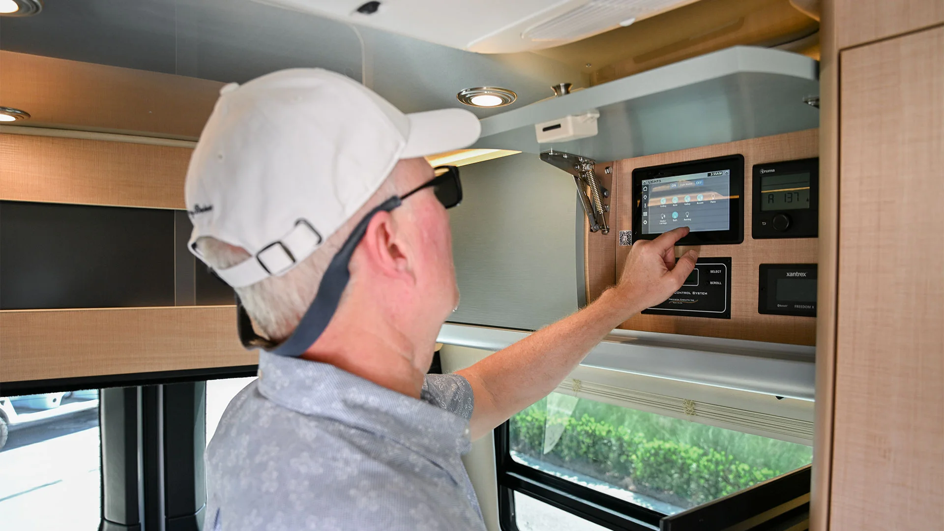 A person in a white cap uses a touchscreen control panel inside an RV, surrounded by modern wooden cabinets and ceiling lights.