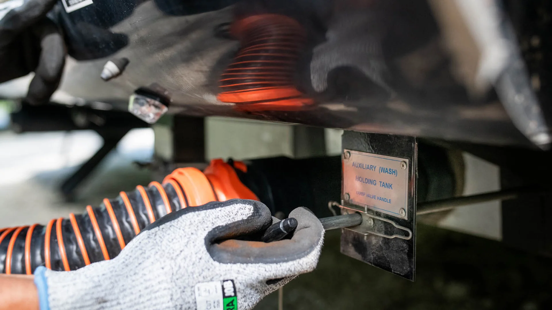 A person wearing a gray work glove connects an orange hose to the auxiliary holding tank of a vehicle, preparing to empty or fill the tank.